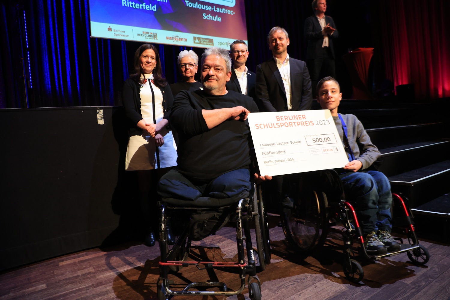 Mehrere Menschen, ganz vorn zwei Männer im Rollstuhl, mit einem Förderpreis-Scheck in Händen.