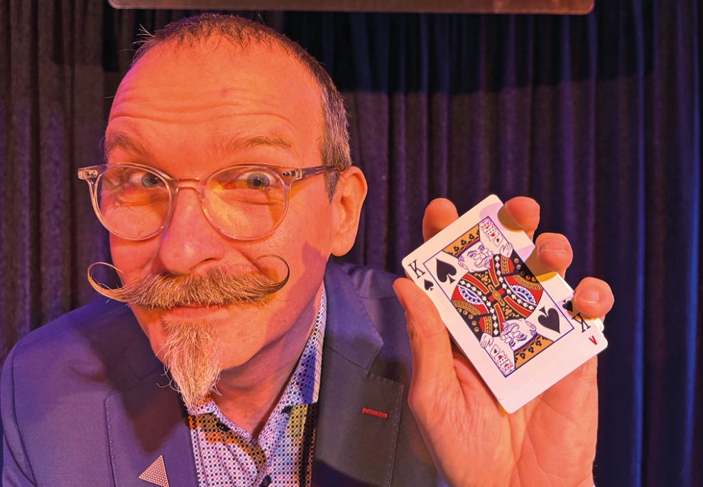 Ein Mann mit Brille und Zwirbelbart schaut in die Kamera und hält eine Spielkarte hoch.