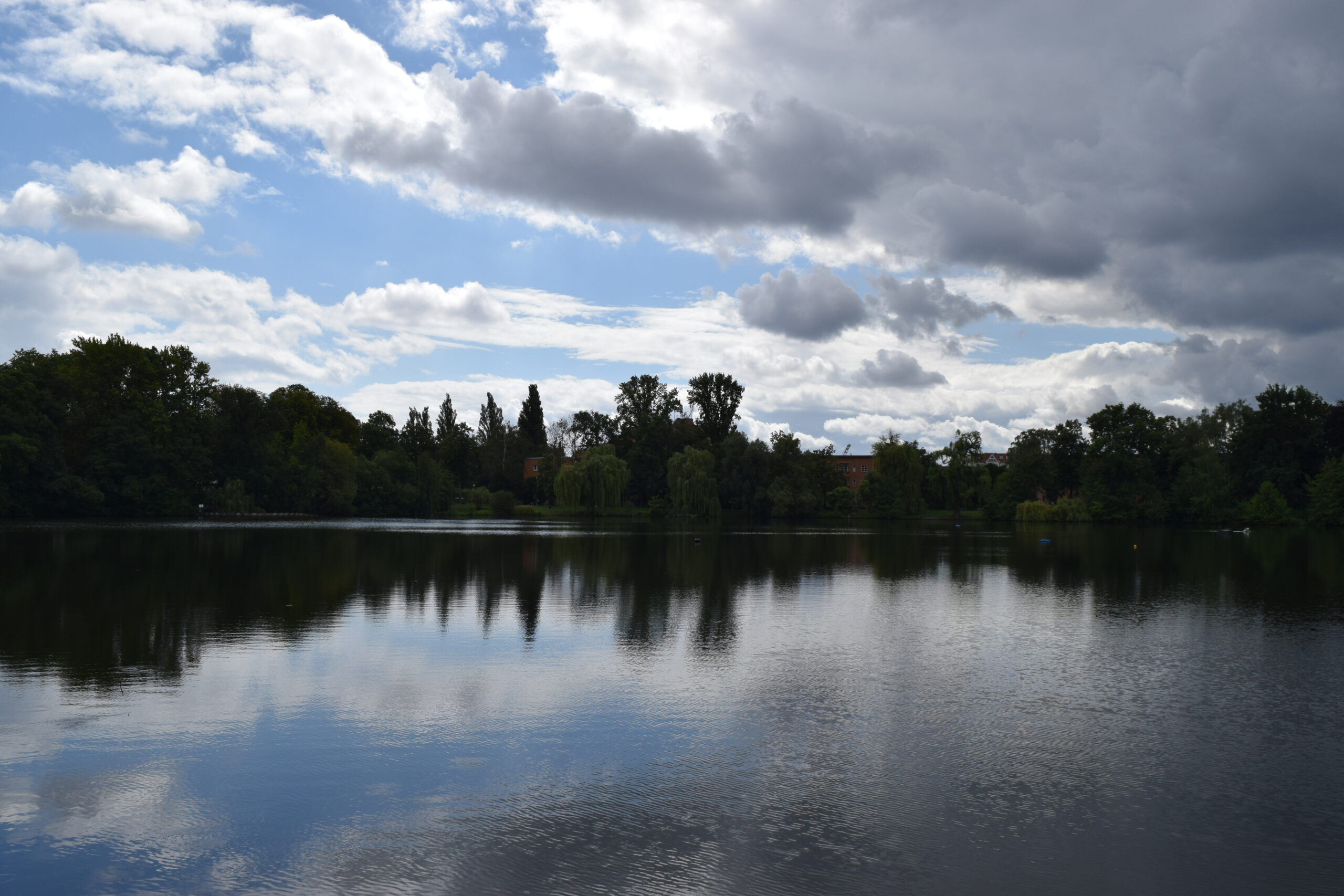 Ein See mit einem fernen Ufer mit Baumbestand. Darüber der halbbewölkte Himmel, der sich im Wasser spiegelt.