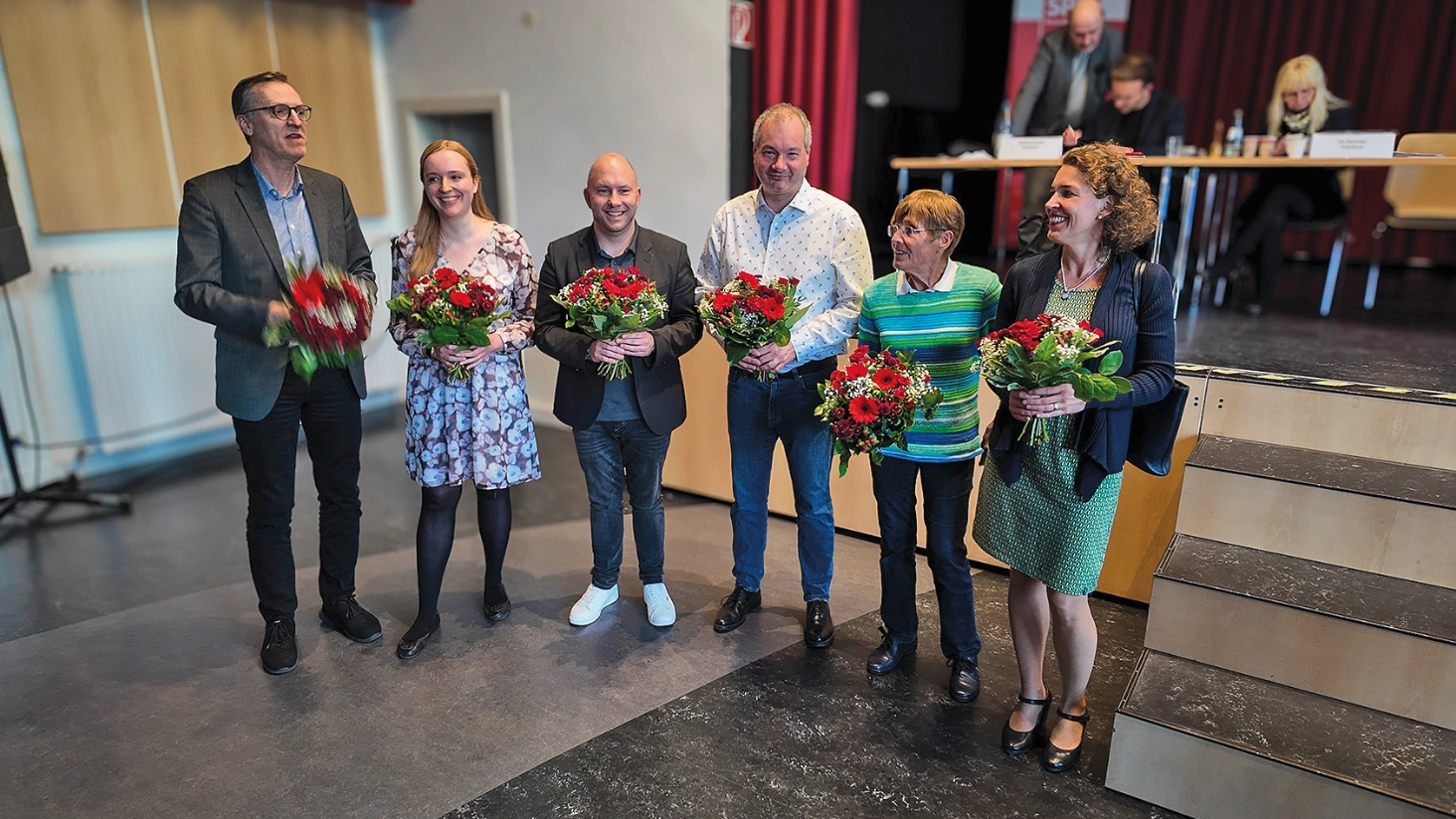 Sechs Menschen mit Blumensträußen vor einer Bühne