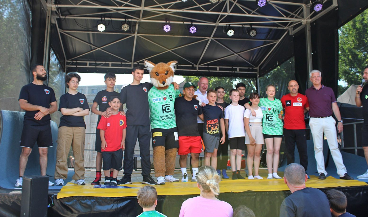 Auf einer Bühne stehen Kinder, Jugendliche und Erwachsene Arm in Arm mit einem Fuchs-Maskottchen.