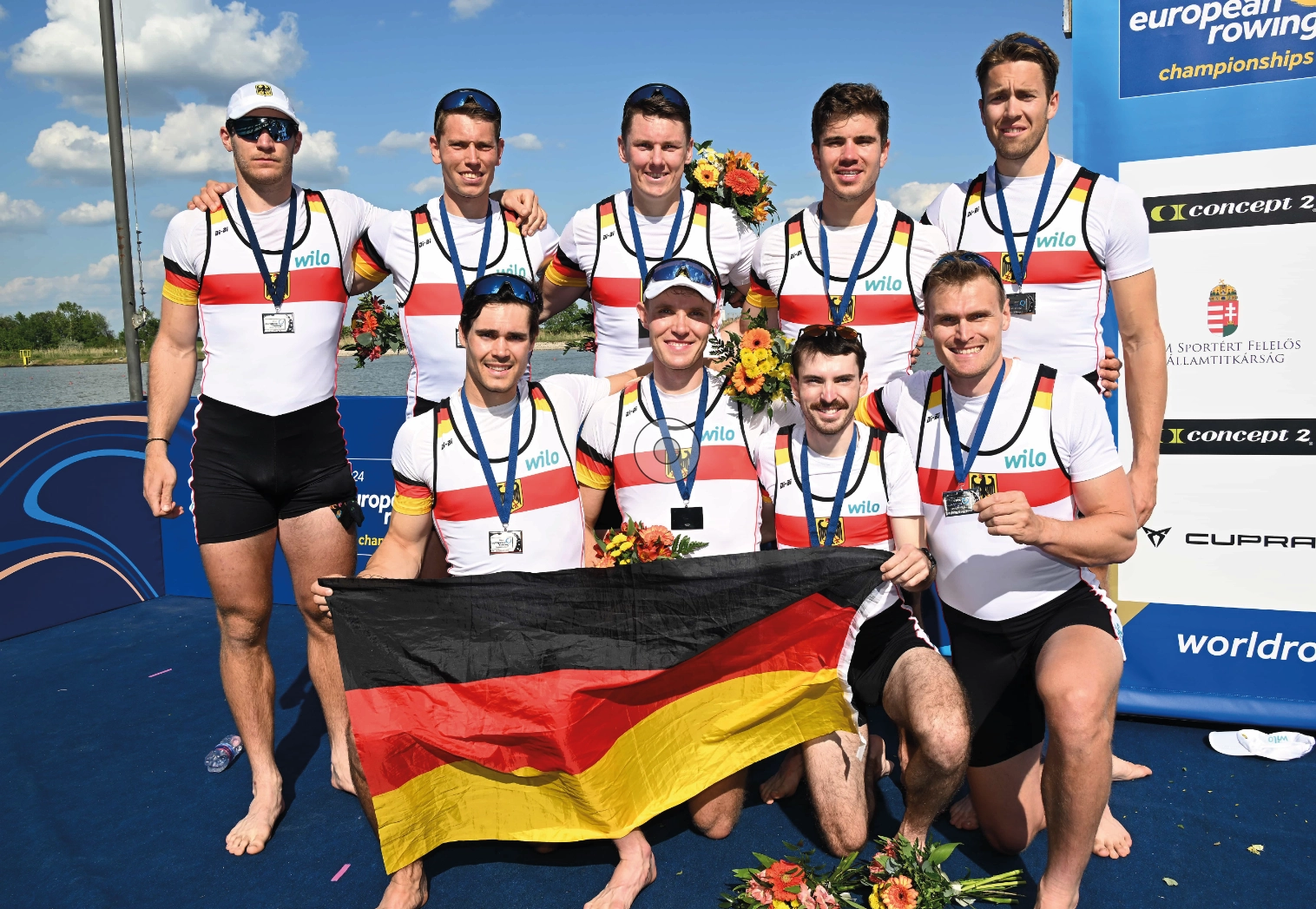 Neun Medaillenträger mit der Deutschlandflagge stehen und knien vor einem Boot.