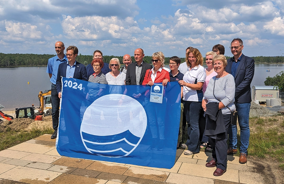 eine große Menschengruppe an einem Seeufer hält gemeinsam eine blaue Flagge mit weiß-blauem Wellen-Logo.