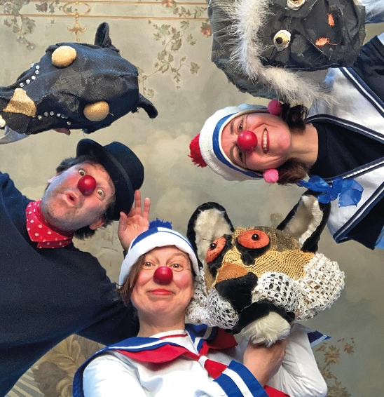 Drei Clowns mit Puppen- und Maskenfiguren lachen in die Kamera