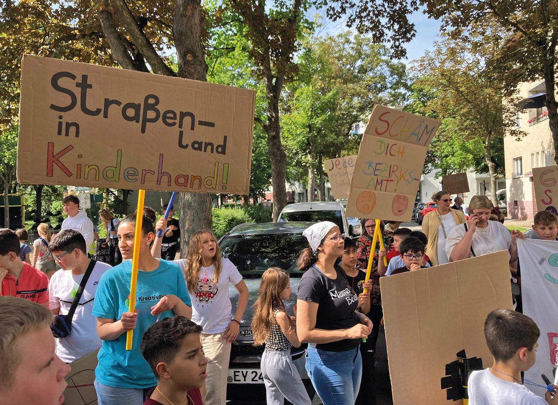 Die Demonstration zeigt eine Gruppe von Menschen, darunter viele Kinder, die auf einer Straße marschieren und Plakate tragen. Ein großes Schild trägt die Aufschrift "Straßen - in Kinderhand!" und eine Frau hält ein Plakat mit der Aufschrift "Schäm dich Bezirksamt." Die Teilnehmer wirken entschlossen und engagiert, und die Umgebung deutet auf eine städtische Straße mit Bäumen im Hintergrund hin.