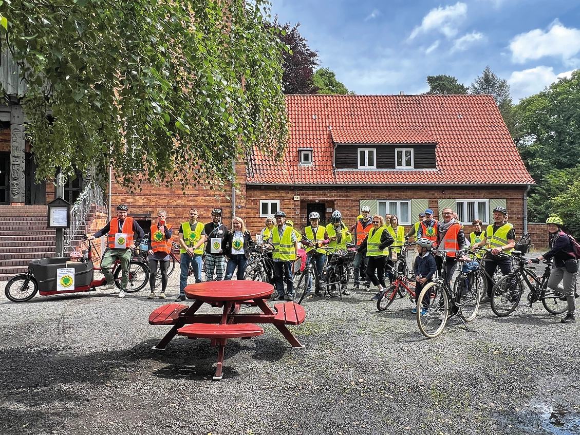 Eine Gruppe von Menschen in Warnwesten hat sich mit Fahrrädern auf einem Platz versammelt. In der Mitte steht ein Tisch mit Bänken.