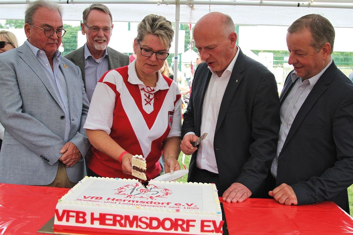 Das Bild zeigt vier Männer und eine Frau, die in der Mitte steht. Sie schneidet eine weiße Torte mit rotem Schriftzug "VFB HERMSDORF EV" an. Die Männer schauen ihr dabei zu.