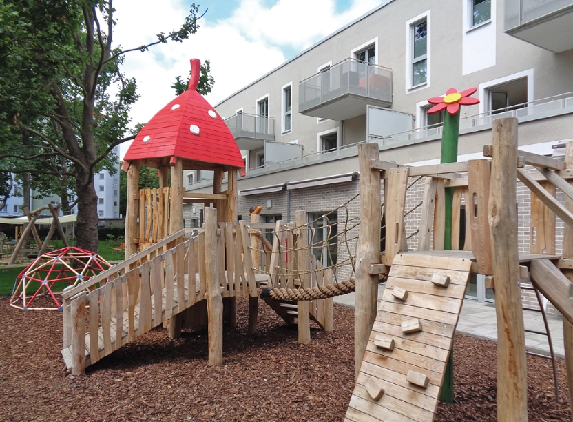 Ein Spielplatz vor einem Gebäude: Klettergerüste in Holzoptik mit einem spitzen Dach in den Farben eines Fliegenpilzes.