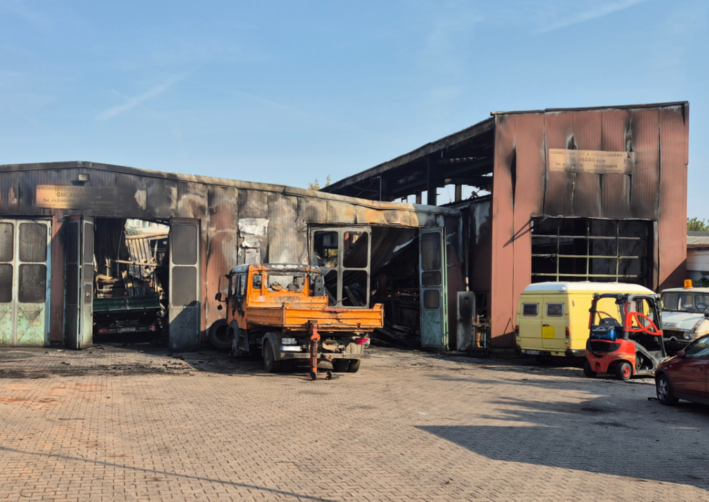 Ein Platz mit Lagerhallen im Hintergrund, davor ein Lkw, alles ist von einem Feuer gezeichnet.