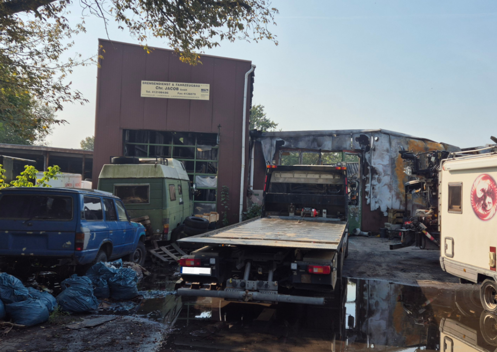 Eine Lagerhalle im Hintergrund, davor mehrere Transportfahrzeuge, alles ist von einem Feuer gezeichnet. Auf dem Boden eine große Löschwasser-Pfütze