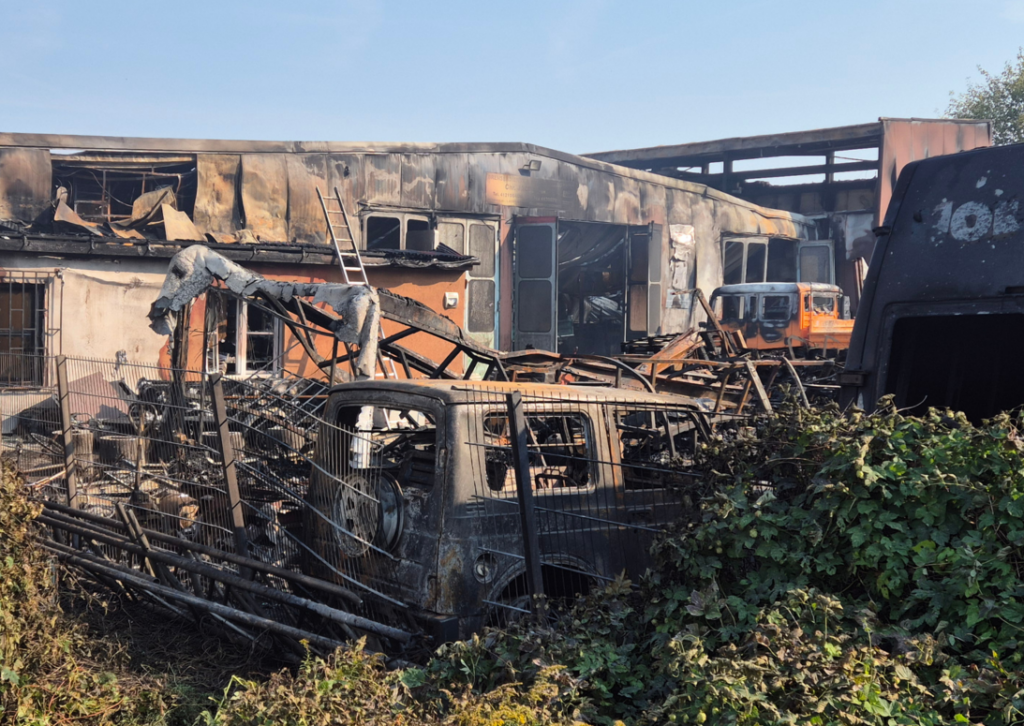 Lagerhallen im Hintergrund, davor ausgebrannte Gerippe von Gebäuden und Fahrzeugen, alles ist von einem Feuer gezeichnet.