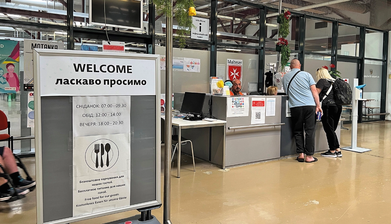 Eine Innenaufnahme des Ankunftszentrums Tegel. Im Vordergrund ein Schild mit einer Willkommensbotschaft in verschiedenen Sprachen, im Hintergrund ein Empfangstresen mit Menschen.