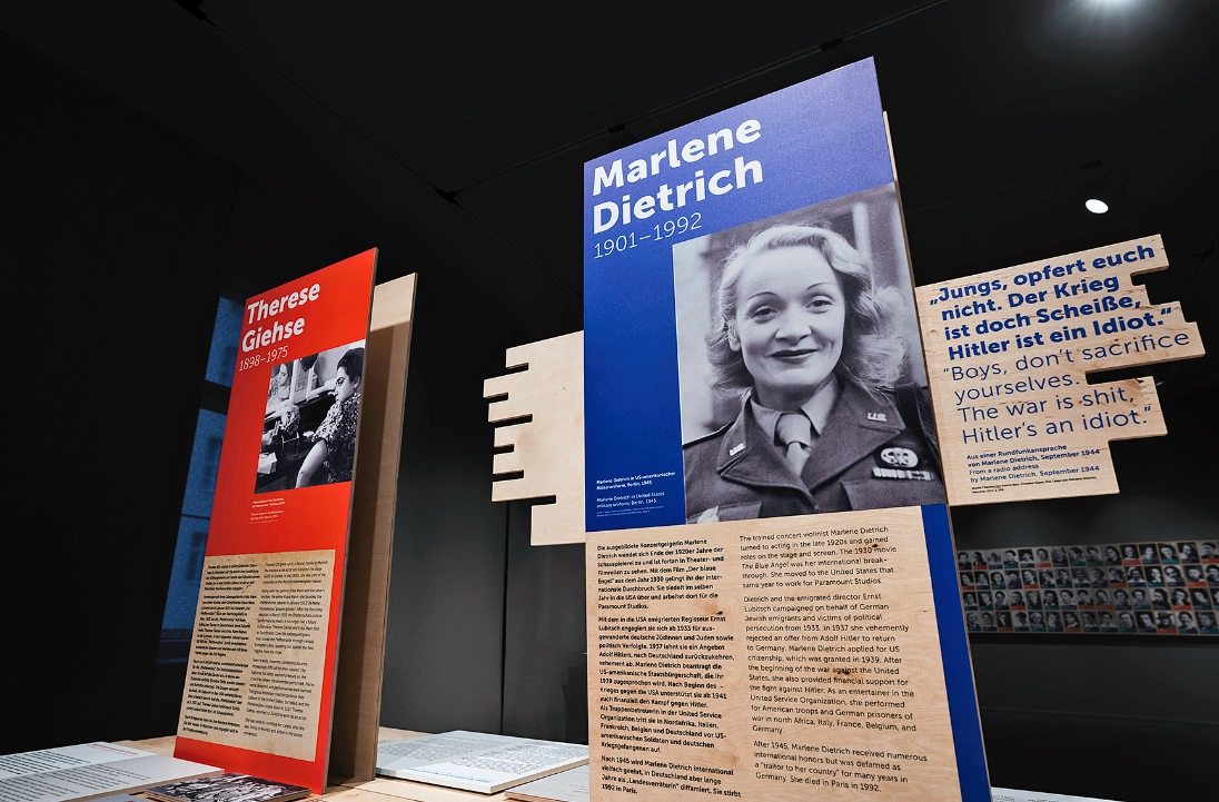 Plakate und Tafeln einer Ausstellung mit großformatigen Fotoaufnahmen und Text. Vorn sieht man einen Aufsteller mit Marlene Dietrich.