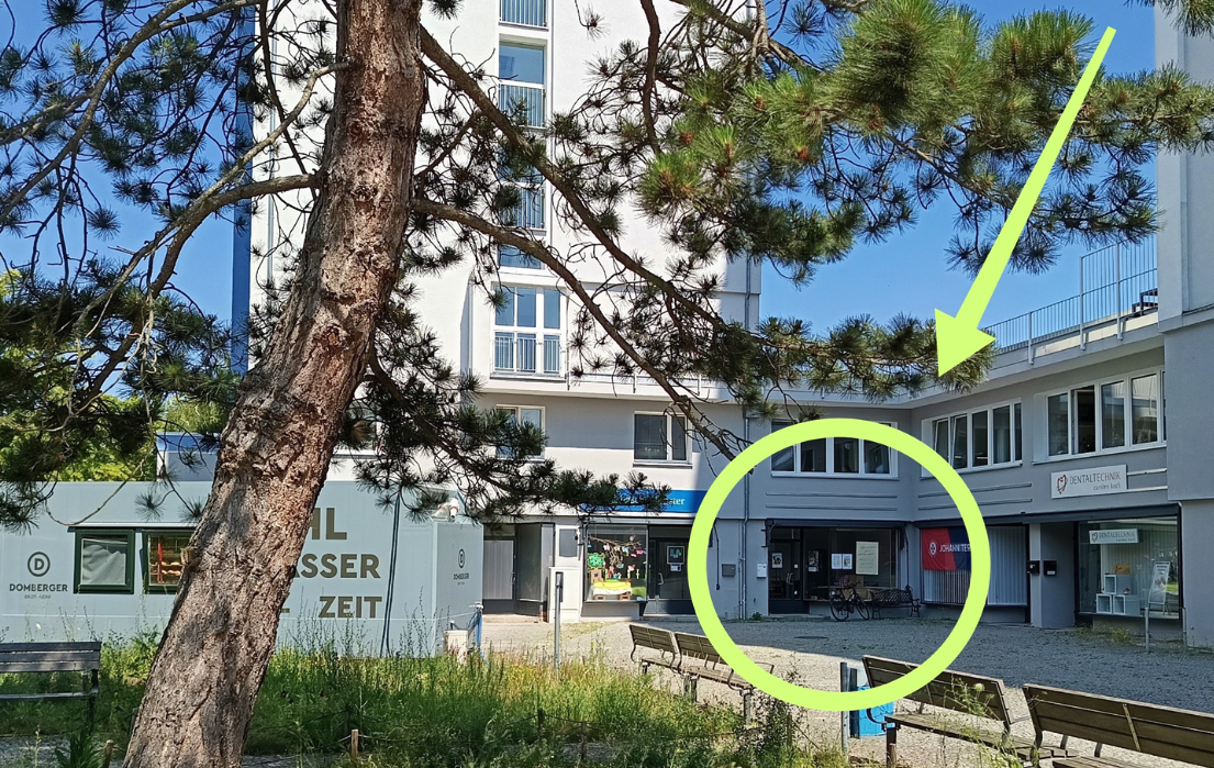 Ein weißes Gebäude vor blauem Himmel, im Vordergrund ein Baum. Ein Teil des Gebäudes ist mit einer gelben Markierung auf dem Foto eingekreist, auf den Kreis deutet ein gelber Pfeil.