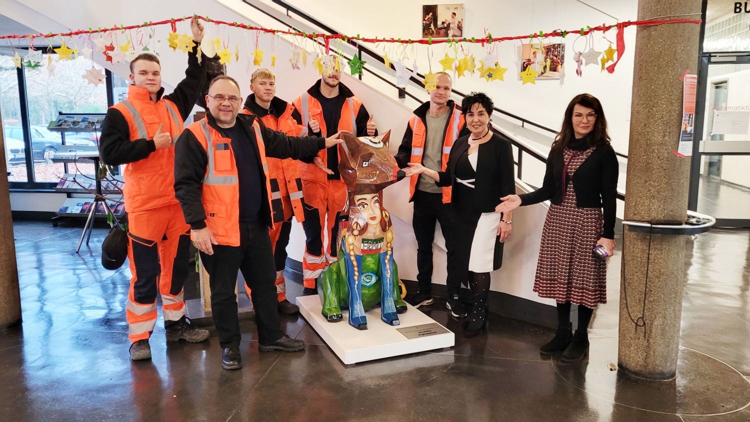 Eine Gruppe Menschen in einem Gebäude-Foyer, viele davon in orangefarbenen Warnwesten. In der Mitte der Gruppe steht eine bunte Fuchs-Skulptur.