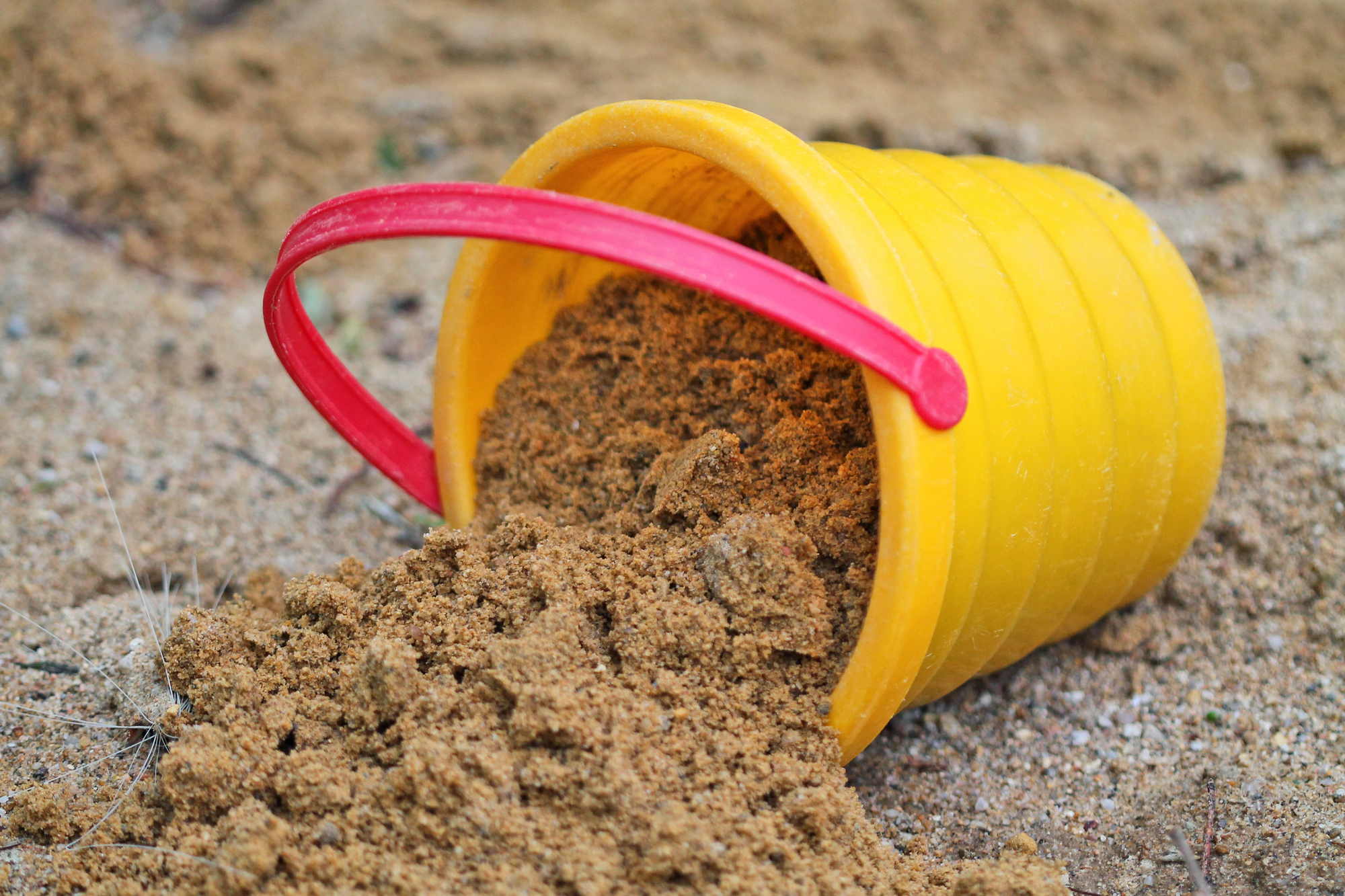 Ein gelber Eimer voller Sand mit einem roten Henkel ist umgekippt, der Inhalt teils herausgekippt.