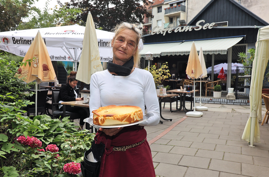 Eine Frau steht mit einem Käsekuchen in Händen auf der Terrasse eines Cafés. Im Hintergrund eingeklappte Sonnenschirme und Stühle.