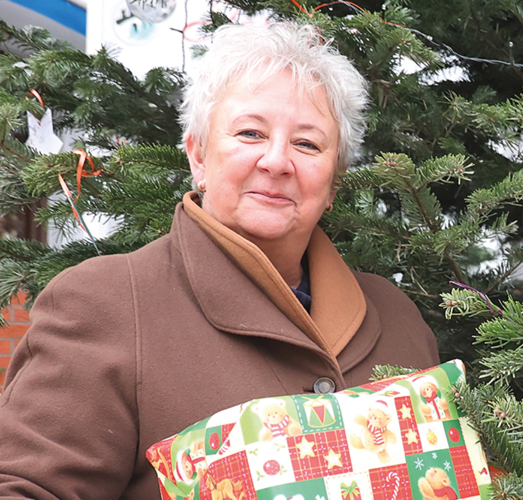 Eine Frau vor einem Nadelbaum. Sie hält ein in Geschenkpapier verpacktes Geschenk in der Hand.