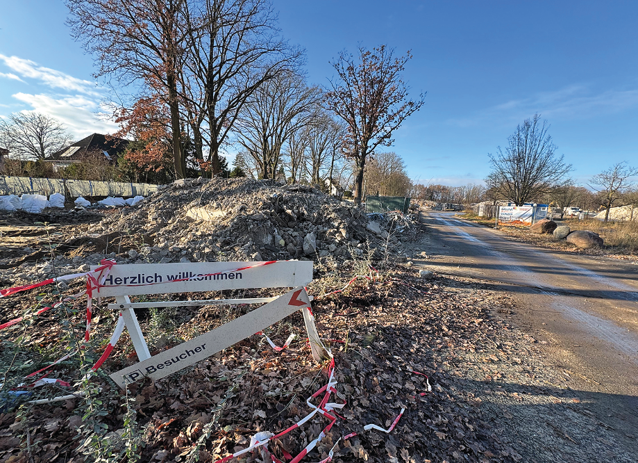 Ein Feldweg und Brachgelände. Im Vordergrund ein kaputtes altes Schild, auf dem noch "herzlich willkommen" zu lesen ist.