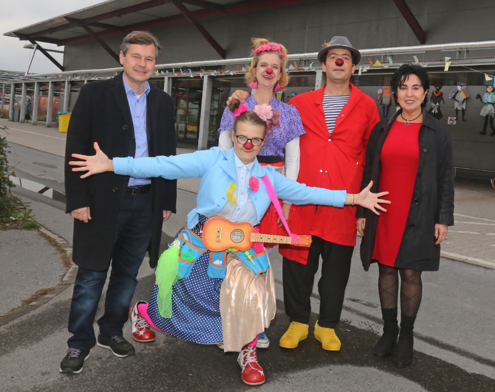 Eine Gruppe von fünf Personen, davon drei Menschen im Clownskostüm. Vorn hält eine Clownin die Arme ausgebreitet; vor ihrer Brust hängt eine Ukulele