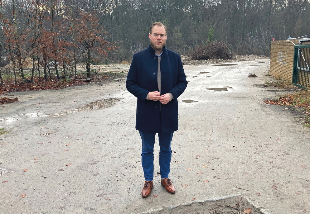 Ein Mann steht auf einem breiten Kiesweg mit vielen Pfützen, im Hintergrund winterlicher Wald, seitlich eine Hecke