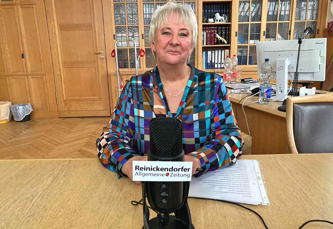 Eine Frau am Schreibtisch eines Arbeitszimmers. Vor ihr auf dem Tisch steht ein Mikrofon mit einem Schild, auf dem "Reinickendorfer Allgemeine Zeitung" steht.