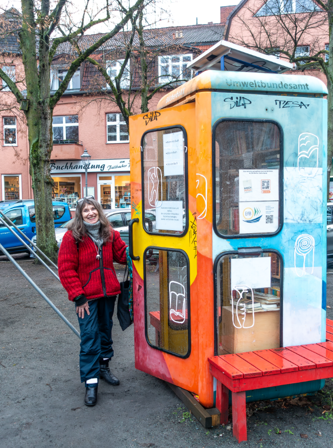 Eine Frau zieht leicht die Tür einer alten, ehemals gelben (und jetzt kunterbunt bemalten) Telefonzelle auf, die eine Büchersammlung beherbergt.