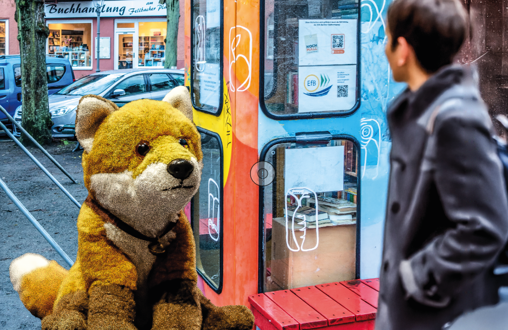 Ein Plüschfuchs im Vordergrund, dahinter eine bunte Telefonzelle, vor der eine Person mit schwarzer Jacke vorübergeht.