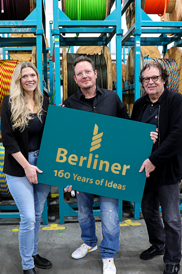 Eine Frau und zwei Männer stehen vor großen Winden mit aufgewickelten bunten Seilen. Sie halten ein Schild mit der Aufschrift "Berliner 160 Years of Ideas".