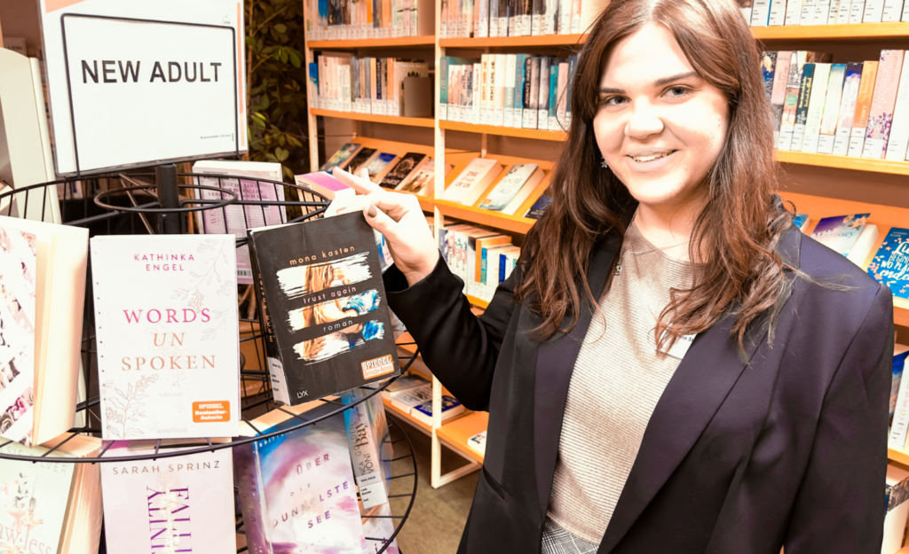 Eine junge Frau in schwarzem Blazer steht in einer Buchhandlung neben einem Aufsteller mit "New Adult" Büchern (entsprechend beschriftet) und zeigt einen Titel in die Kamera.