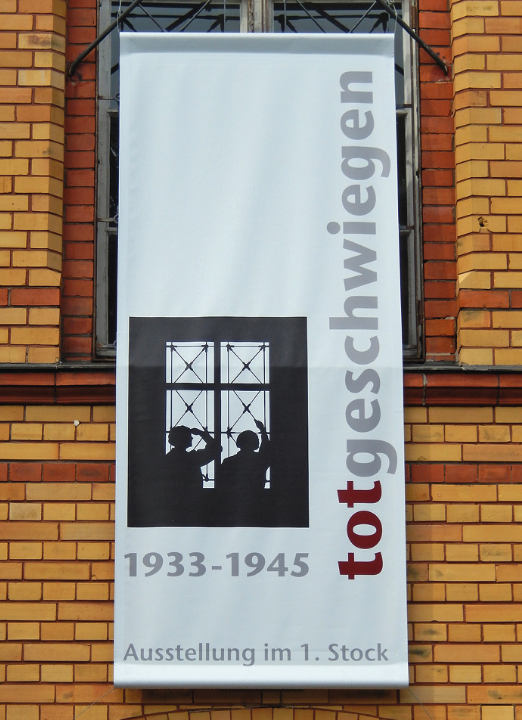 Ein Banner am Fenster eines Gebäudes: Es weist auf die Ausstellung "totgeschwiegen" hin, die im ersten Stock zu sehen ist.