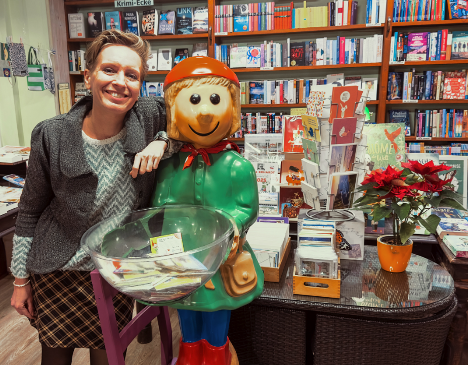 Eine Frau in einer Buchhandlung, im Hintergrund viele Regale vollder Bücher. Ihren Arm lehnt sie auf eine lustige Figur, die eine Schale mit Pixibüchern vorhält.