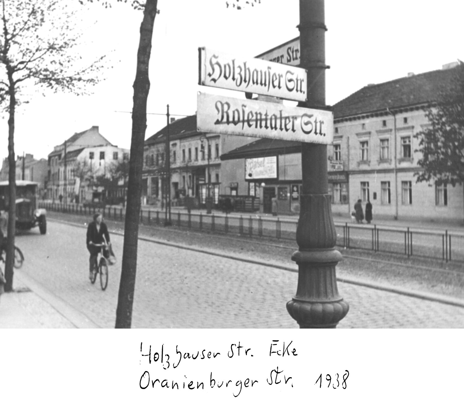 eine historische Fotografie (schwarz/weiß) zeigt einen Pfeiler mit Straßenschildern. Man sieht die durchgestrichene Bezeichnung "Rosenthaler Straße", darüber "Holzhauser Straße".