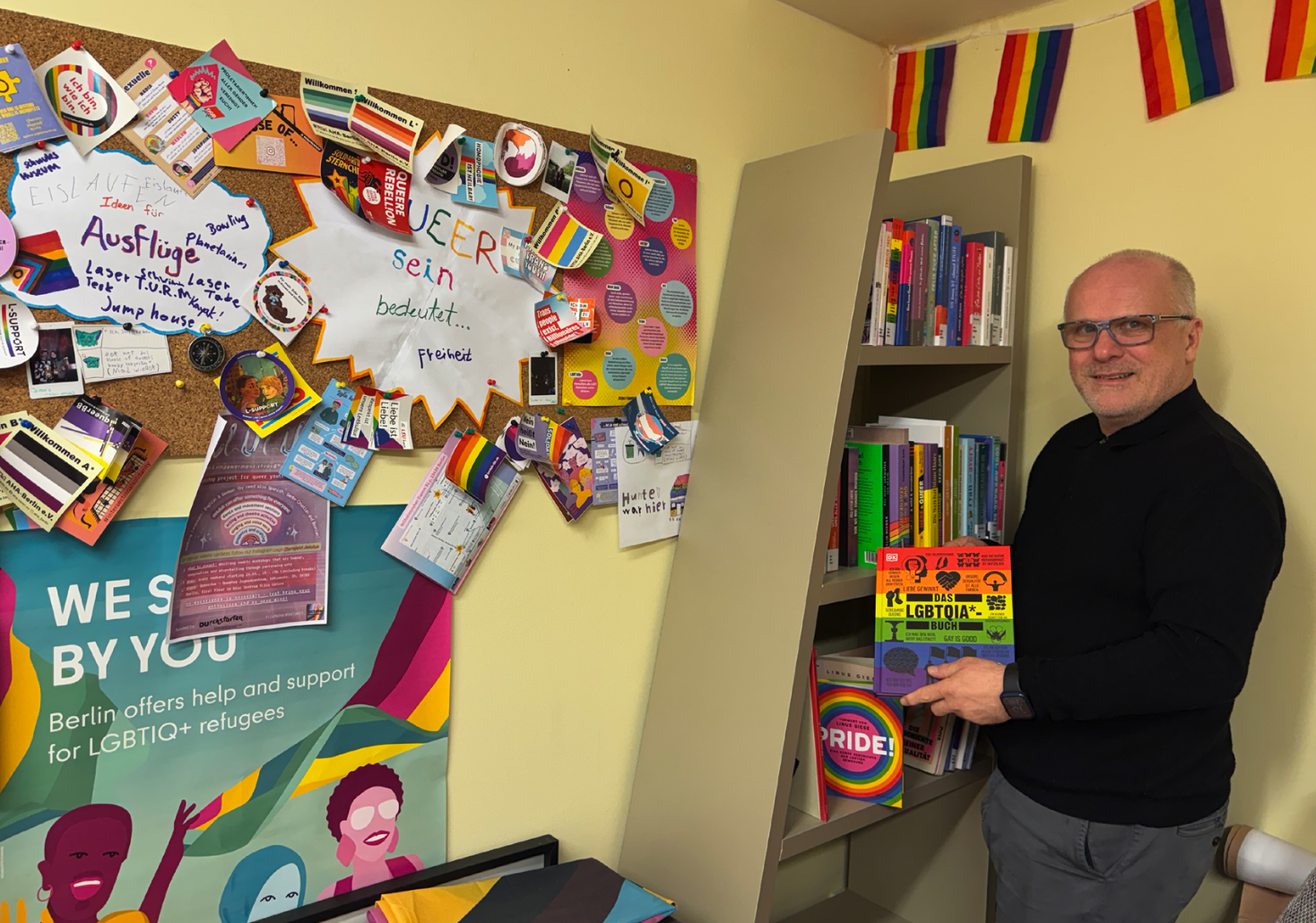 Ein Mann mit einem regenbogenfarbenen Buch in der Hand steht in einem Raum mit Regenbogen-Wimpeln, bunten Büchern im Regal und einer sehr vollen, bunten Pinnwand.