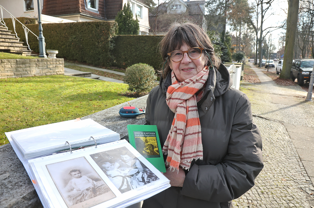 Eine Frau in dicker Jacke und mit Schal steht in einer Straße. In den Händen hält sie einen Ordner mit Abbildungen.