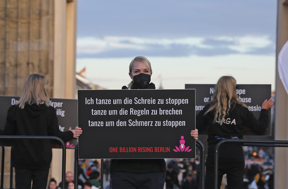 Eine junge Frau mit Maske über Mund und Nase hält unter freiem Himmel ein großes Plakat mit beiden Händen vor ihrem Oberkörper, darauf eine Bekundung gegen Gewalt an Frauen.