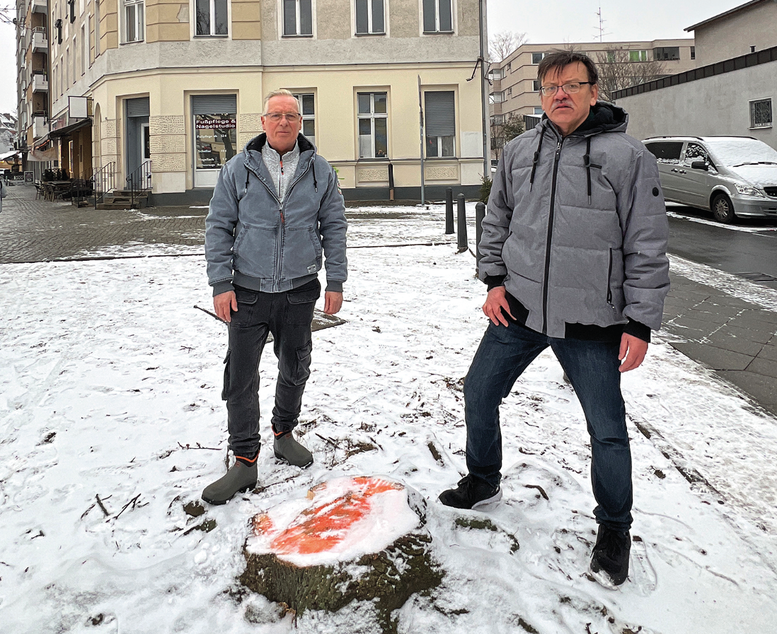 Zwei Männer stehen in winterlichem Wetter mit Schnee am Boden vor einem Baumstumpf, der frisch abgesägt ist.