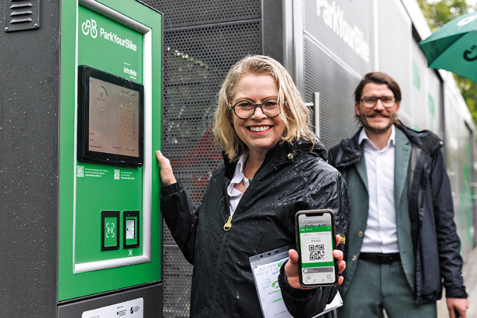 Eine Frau und ein Mann stehen vor einem grünen Kasten. Die Fraut hält eine App in die Kamera.
