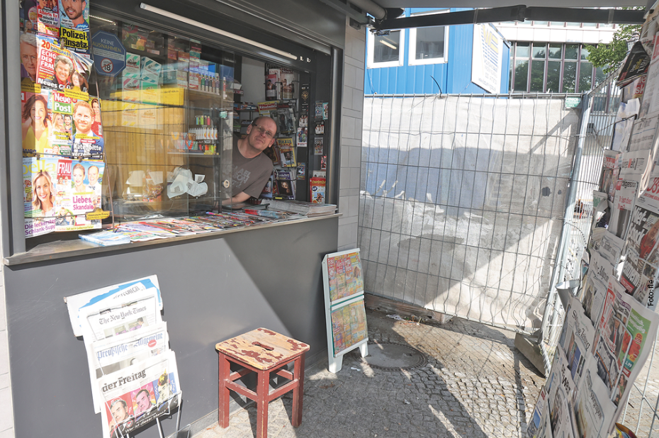 Ein Mann schaut aus einem Kioskfenster. Der Kiosk ist von einem Bauzaun umgeben.