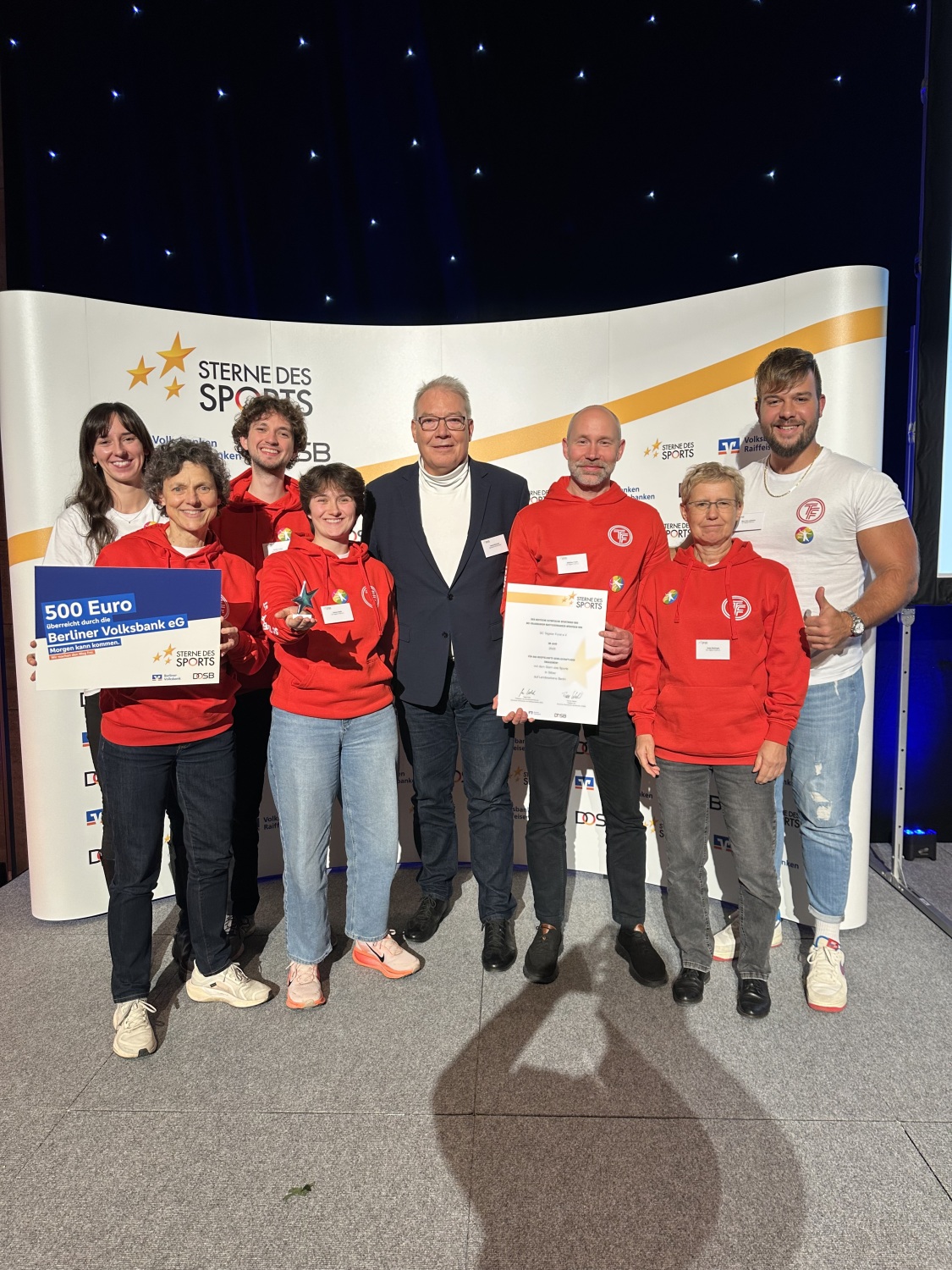 Eine Gruppe Menschen mit Urkunden vor einer Fotowand, auf der "Sterne des Sports" steht.