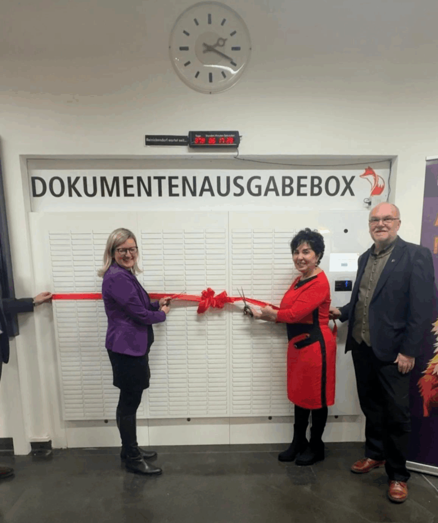 Zwei Frauen stehen vor der Dokumentenausgabebox und sind im Begriff, eine rote Schleife, die um diese gelegt ist, zu durchtrennen. Daneben steht ein Mann.
