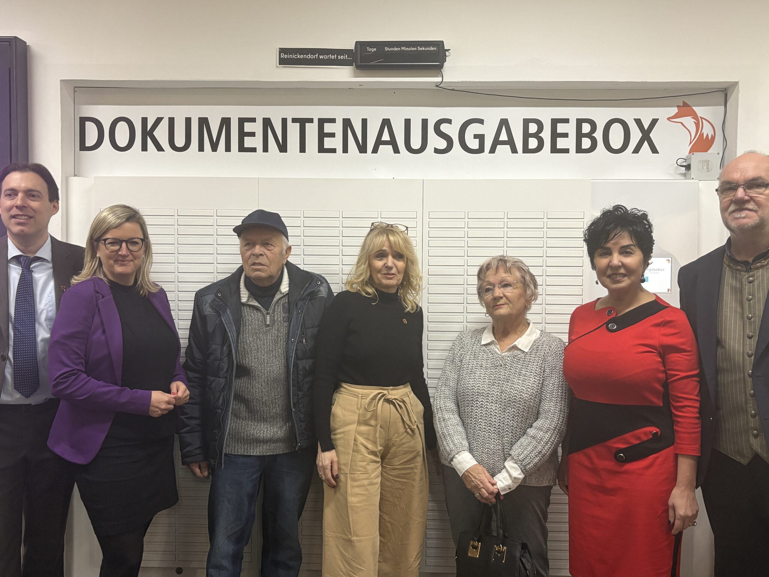 Sieben Menschen stehen vor einem wandfüllenden Gerät, das mit "Dokumentenausgabebox" beschriftet ist.