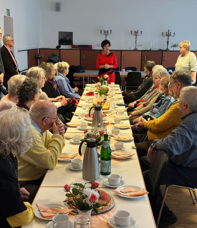 Eine lange Tafel mit Geschirr, daran sitzen zu beiden Seiten viele Menschen. Am Kopf steht eine Frau und spricht zu den anderen.