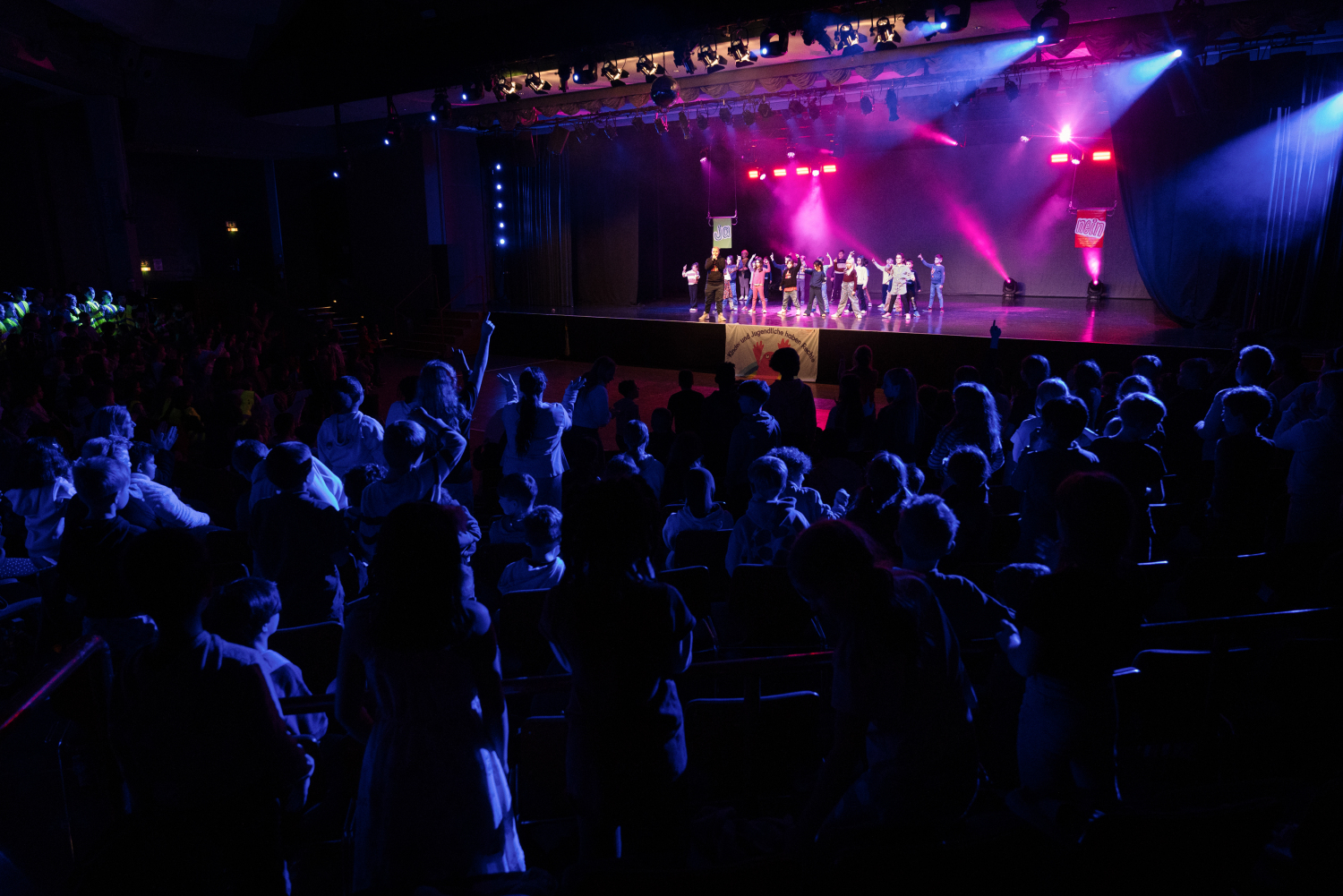 Blick auf eine lila beleuchtete Bühne in einem dunklen Saal. Die Perspektive geht über das Publikum hinweg. Auf der Bühne tanzen Kinder.