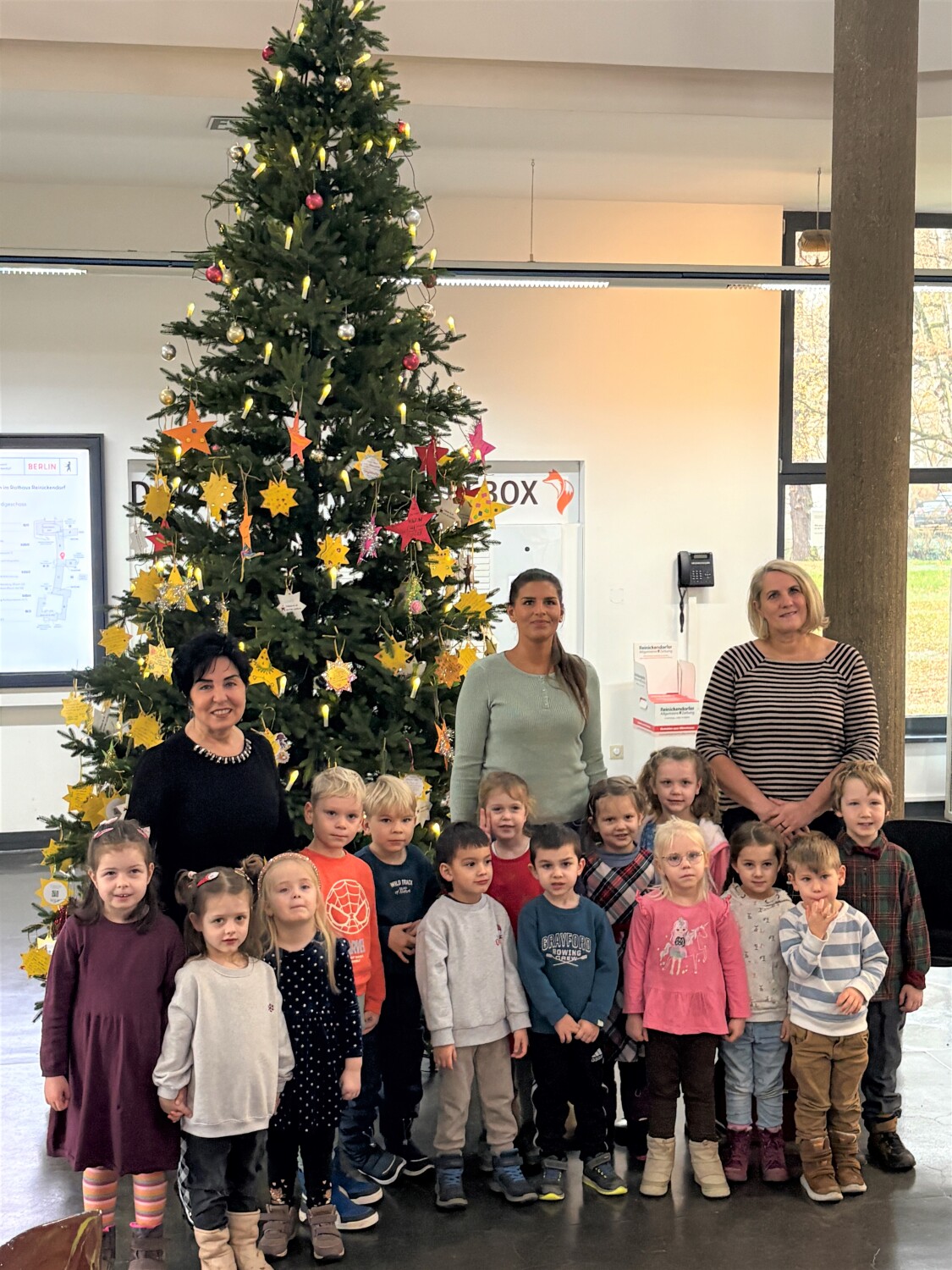 Eine Gruppe Kinder und drei erwachsene Frauen vor einem geschmückten Weihnachtsbaum in einem großen, hohen Innenraum