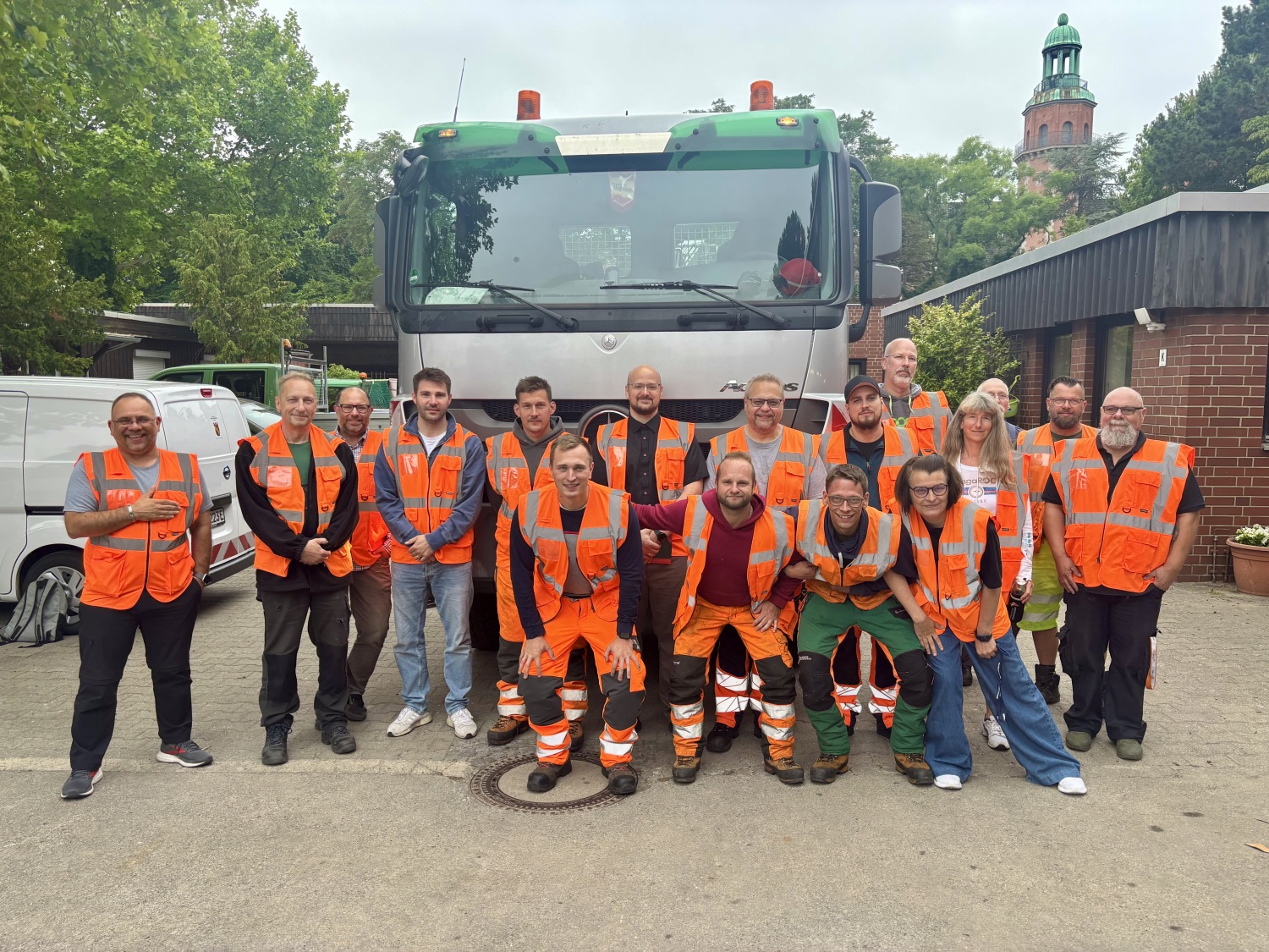Eine Gruppe von Menschen mit orangefarbenen Warnwesten steht und kniet für ein Gruppenfoto vor einem Lastwagen.