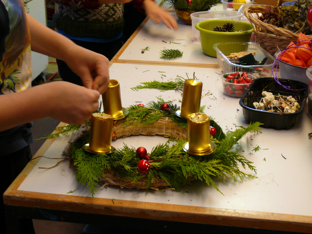 Auf einem Tisch wird gebastelt: Im Vordergrund sieht man einen Adventskranz, fast fertig, mit vier goldnenen Kerzen