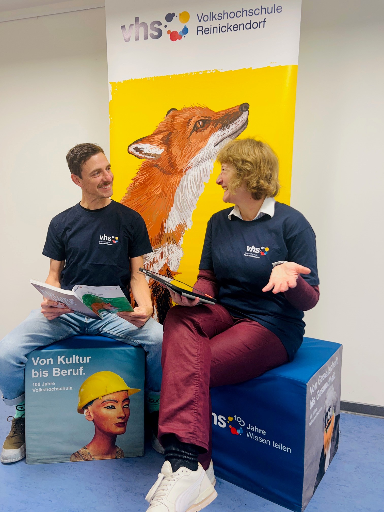 Zwei Menschen sitzend vor einem Plakat mit einem Fuchs darauf