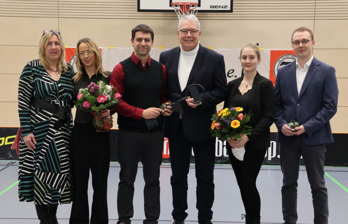 Eine Gruppe von sechs Personen, aufgestellt zu einem Foto, in einer Sporthalle. Zwei von ihnen halten Blumensträuße im Arm.