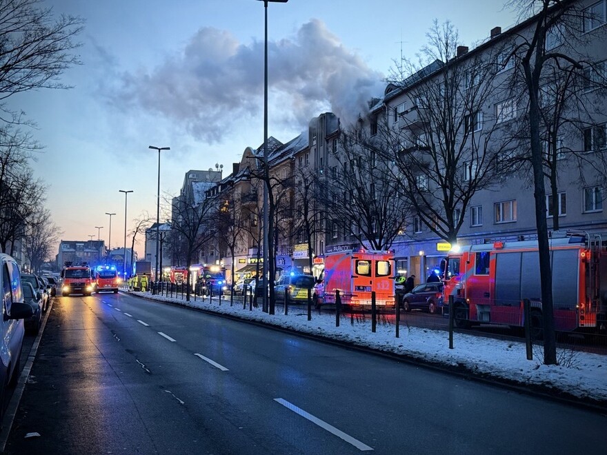 Ein Straßenzug in einer Stadt, keine Autos, nur Rettungsfahrzeuge sind darauf zu sehen. Aus dem oberen Stock einer Hausfassade dringt Rauch.